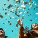 Pareja joven celebrando mientras llueven billetes de dólar y monedas sobre un fondo azul brillante – Mundo Chido