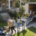 Cinco jóvenes trasladan cajas y muebles de un camión rotulado "Mudanzas Express" a una casa con un cartel de Se Alquila, subiendo las pertenencias por la pasarela y la rampa en un día soleado.